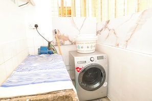 A laundry area with a washing machine, a bed with a striped bedspread, and a window with blinds. - Cozy, comfortable and child friendly (Nairobi)