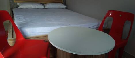 A single bed with a wooden headboard, two red plastic chairs, and a small round table in a room with plain walls.