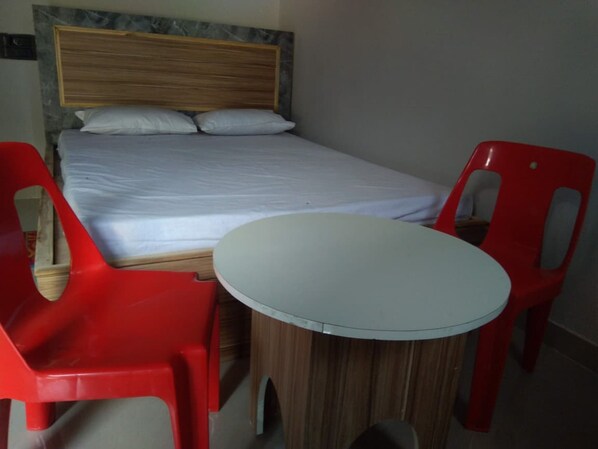 Una cama individual con cabecera de madera, dos sillas de plástico rojo y una mesita redonda en una habitación con paredes lisas.