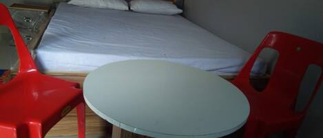 A bed with a wooden headboard, two pillows, and a white mattress. There is a small round table with a white top and two red chairs.