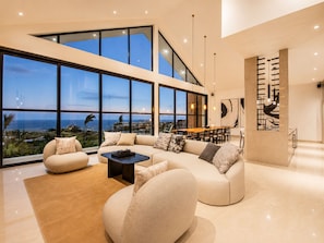 A modern living room with a large window, a sofa set, a coffee table, and a dining area. - Ocean View Mansion Sauna, Cold Plunge & Gym (Nusa Dua)