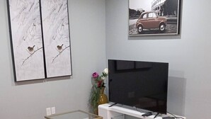 Un salon moderne avec un canapé en cuir noir, une table basse en verre, un téléviseur à écran plat posé sur un meuble blanc, et deux tableaux encadrés accrochés au mur.