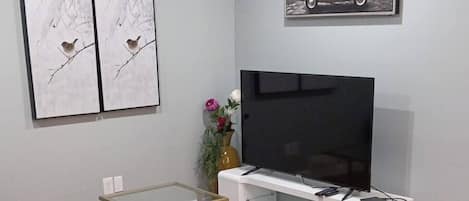 A modern living room with a black leather sofa, a glass coffee table, a flat-screen TV on a white stand, and two framed pictures on the wall.