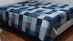 Un lit simple avec une couvre-lit à motifs, dans une pièce au plancher en bois et dotée d’une tête de lit blanche.