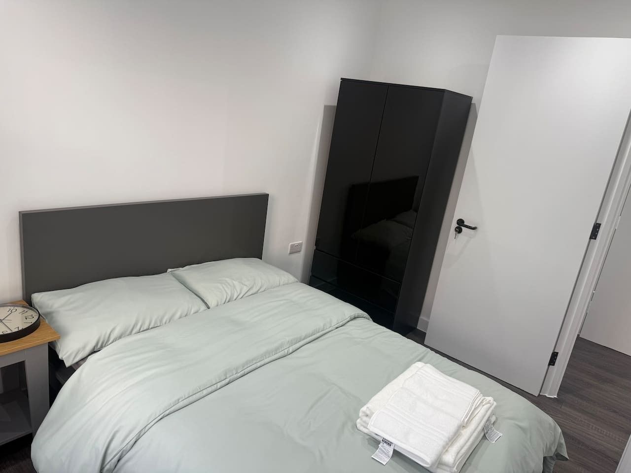 A single bed with a grey headboard, a bedside table with a clock, and a wardrobe.