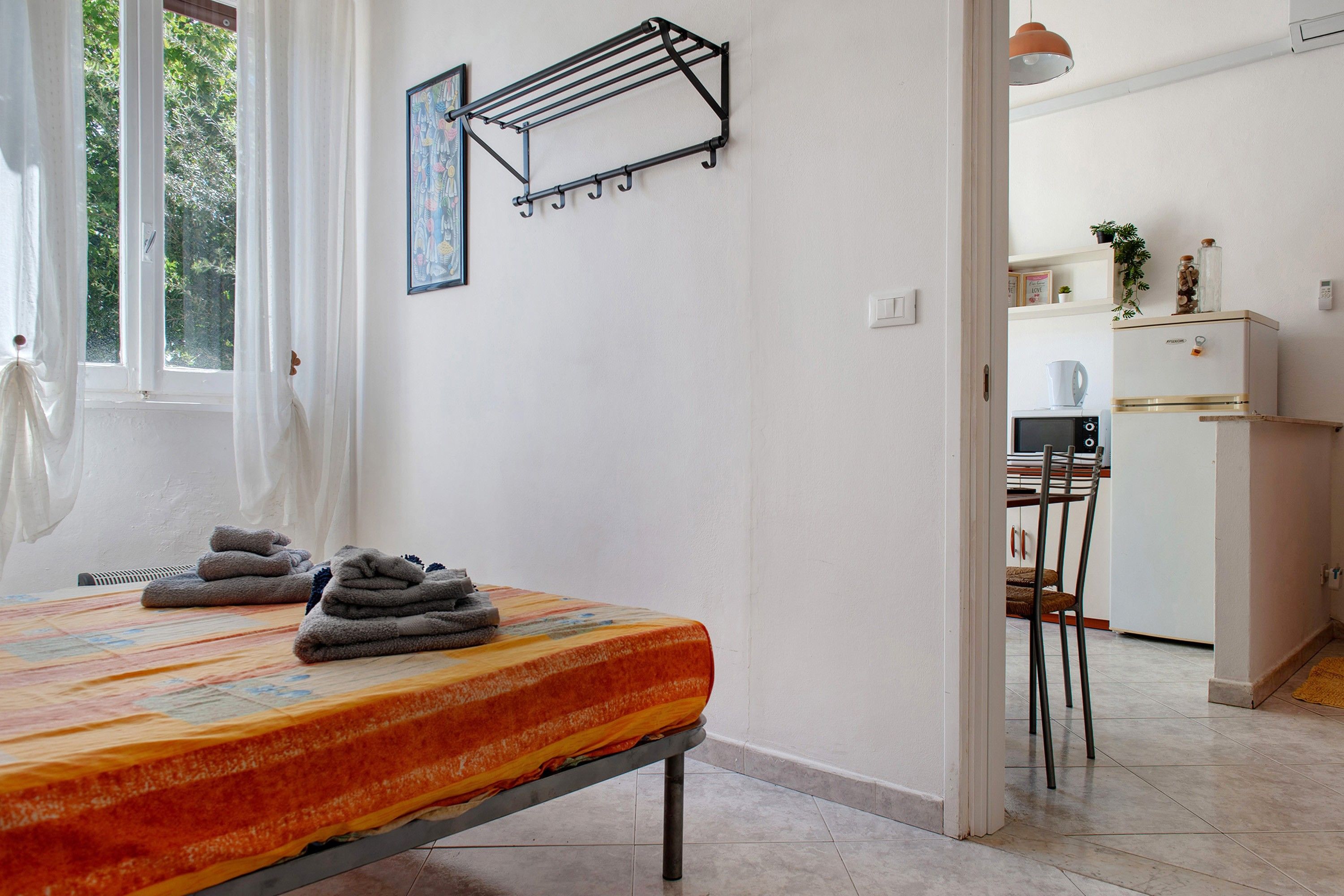 A bedroom with a bed, a drying rack, a window with curtains, and a view into the kitchen area.
