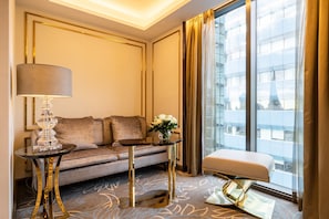 A modern hotel room with a sofa, a side table, a lamp, and a view of a building through large windows.