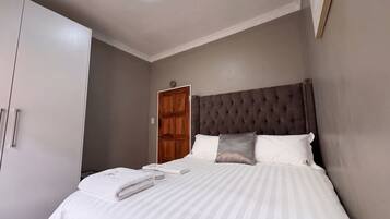 Een net opgemaakt bed met kussens en een opgevouwen handdoek, een houten garderobe en een gesloten deur in een hotelkamer.