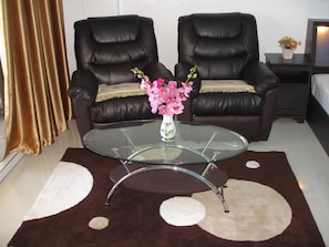 A room with two leather armchairs, a glass coffee table, a vase with flowers, a small bedside table with a plant, and a bed with white bedding.