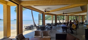 Een overdekt terras met tafels en stoelen, met uitzicht op een strand en de oceaan.