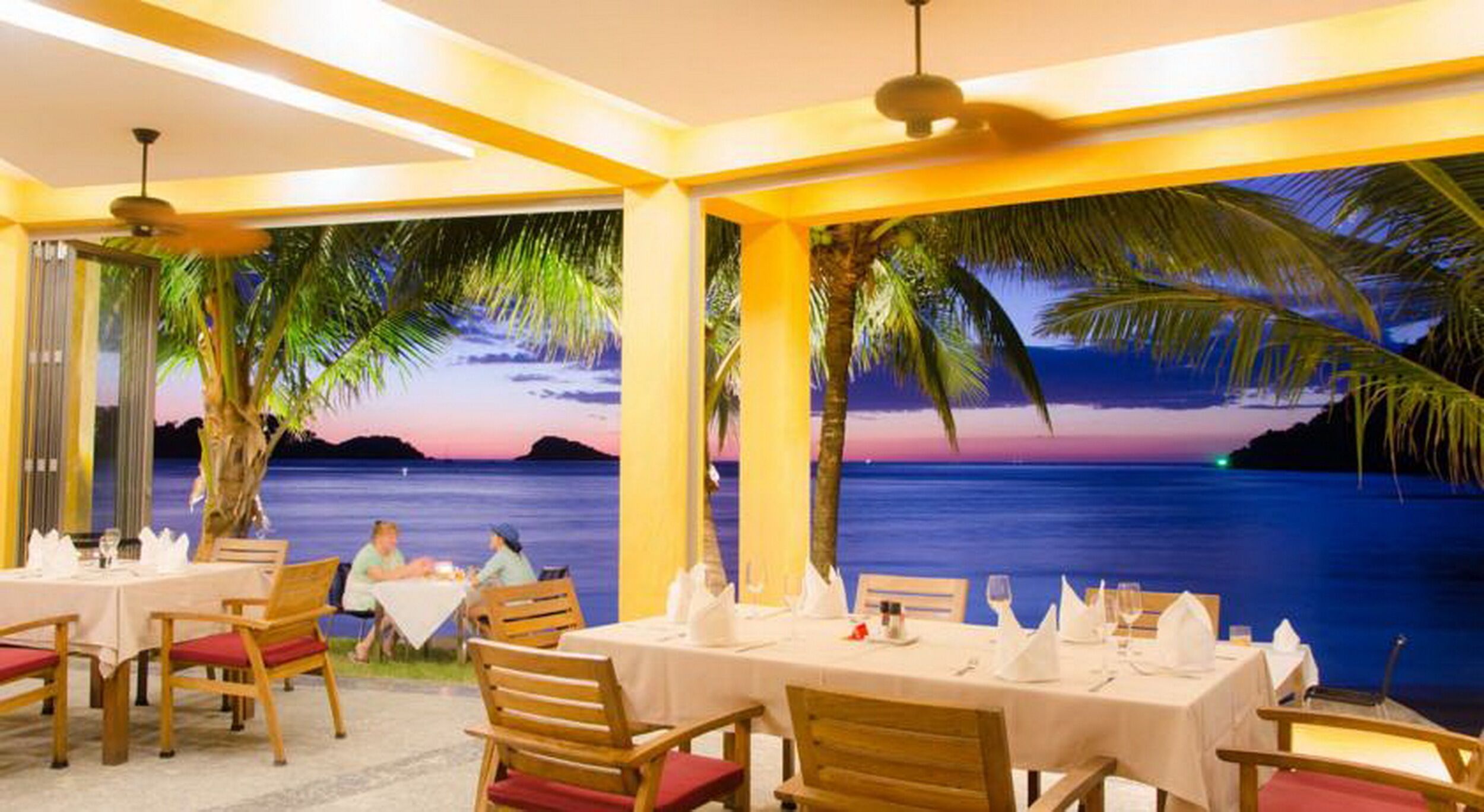 Restaurante com mesas preparadas para refeições, com vista panorâmica do oceano e palmeiras.