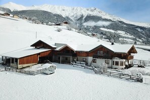 Een chaletachtige constructie met een sneeuwbedekte dak, omringd door een houten omheining, met op de achtergrond besneeuwde bergen.