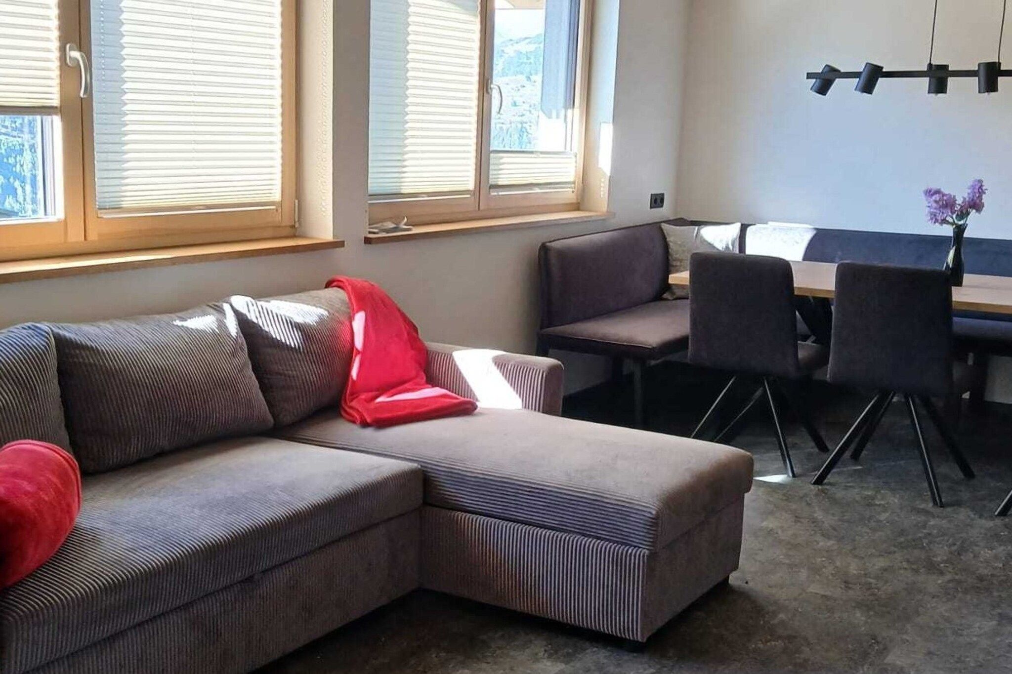 A living room with a sectional sofa, a dining table with chairs, and large windows with blinds.