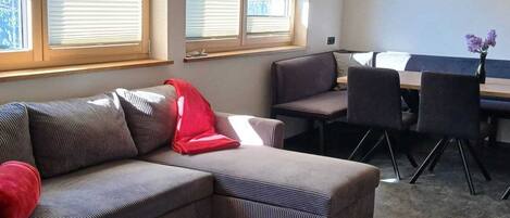 A living room with a sectional sofa, a dining table with chairs, and large windows with blinds.