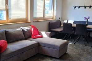 Ein Wohnzimmer mit einem Ecksofa, einem Esstisch mit Stühlen und großen Fenstern mit Jalousien.