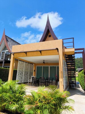A modern building with a triangular roof, a covered patio area with outdoor furniture, and a staircase leading to an upper level. - 4 Bedroom Deluxe Beach Villa 21C, Blue Haven Bay Resort (Ko Chang)