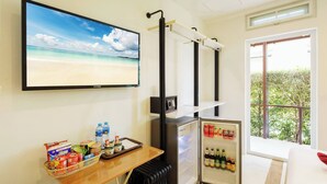 A room with a television displaying a beach scene, a small refrigerator stocked with beverages, a table with snacks, and a window with a view of greenery. - Beach Villa 21D Blue Haven Bay Resort - 4 rooms (Ko Chang)