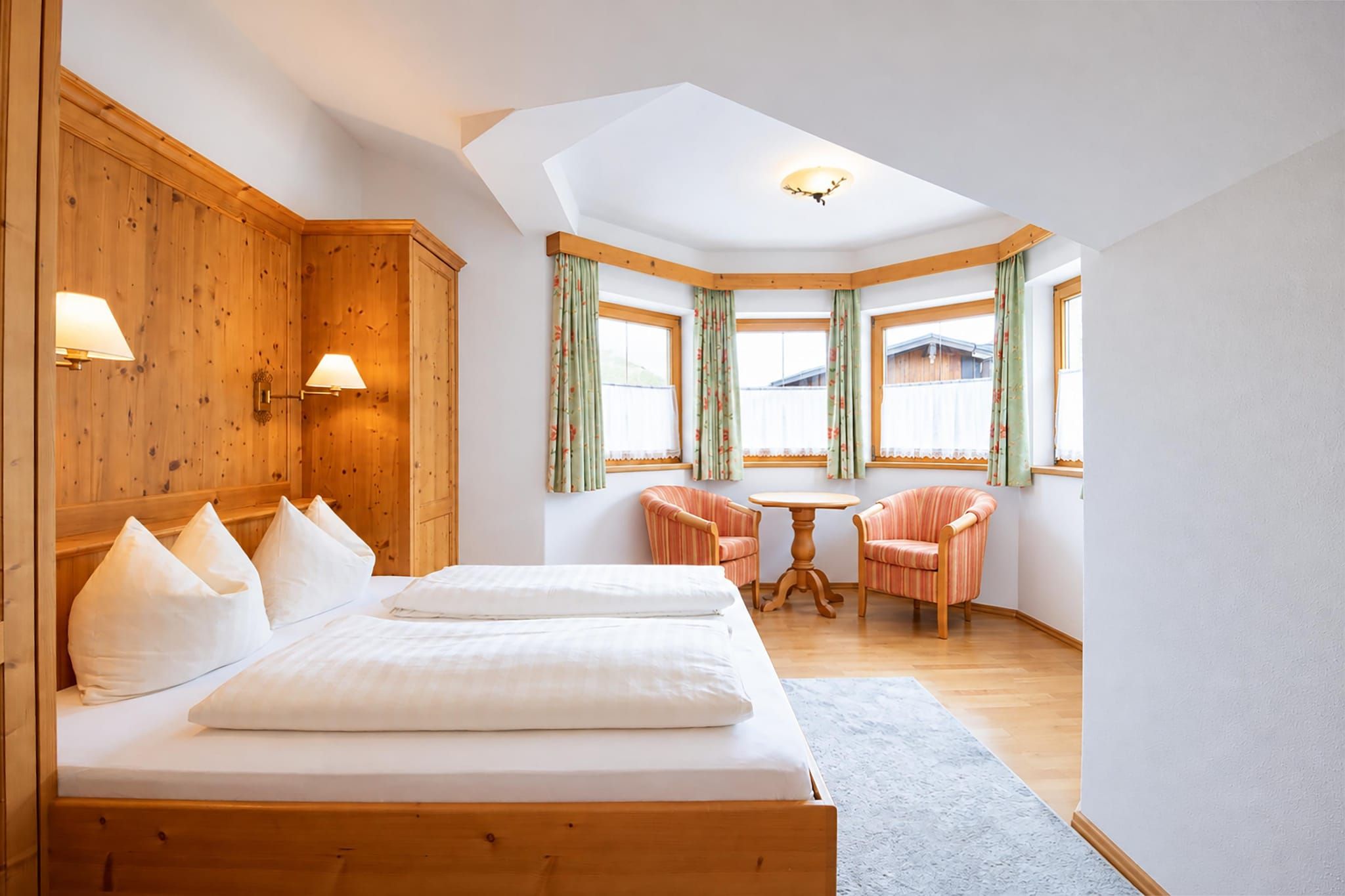 Een slaapkamer met een groot bed, twee stoelen, een klein tafeltje, houten wanden en een raam met gordijnen.