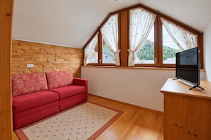 A small room with a red sofa, a wooden wall, a window with curtains, and a television on a wooden table. - Holiday apartment in an Alpine setting (Achenkirch)