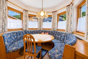 Una mesa redonda con una silla de madera en una habitación de esquina con un banco azul estampado, ventanas amplias con cortinas y una lámpara colgante.