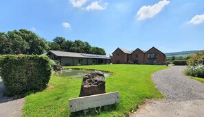 A Victorian barn with a paved path leading to it, surrounded by lush greenery and a small pond. - Osborne Collection at The Victorian Barn Dorset – Sleeps 24, 2 Hot Tubs (Blandford Forum)
