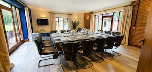A dining area with a long table set for a meal, surrounded by chairs, with a television on the wall and large windows. - Osborne Collection at The Victorian Barn Dorset – Sleeps 24, 2 Hot Tubs (Blandford Forum)