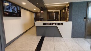 A reception area with a reception desk, a flat-screen TV mounted on the wall, and a patterned wall to the right. - HOTEL HILL VIEW (Ahmedabad)