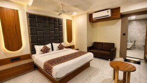 A modern bedroom with a large bed, a ceiling fan, an air conditioning unit, a seating area, and a view of the bathroom. - HOTEL HILL VIEW (Ahmedabad)