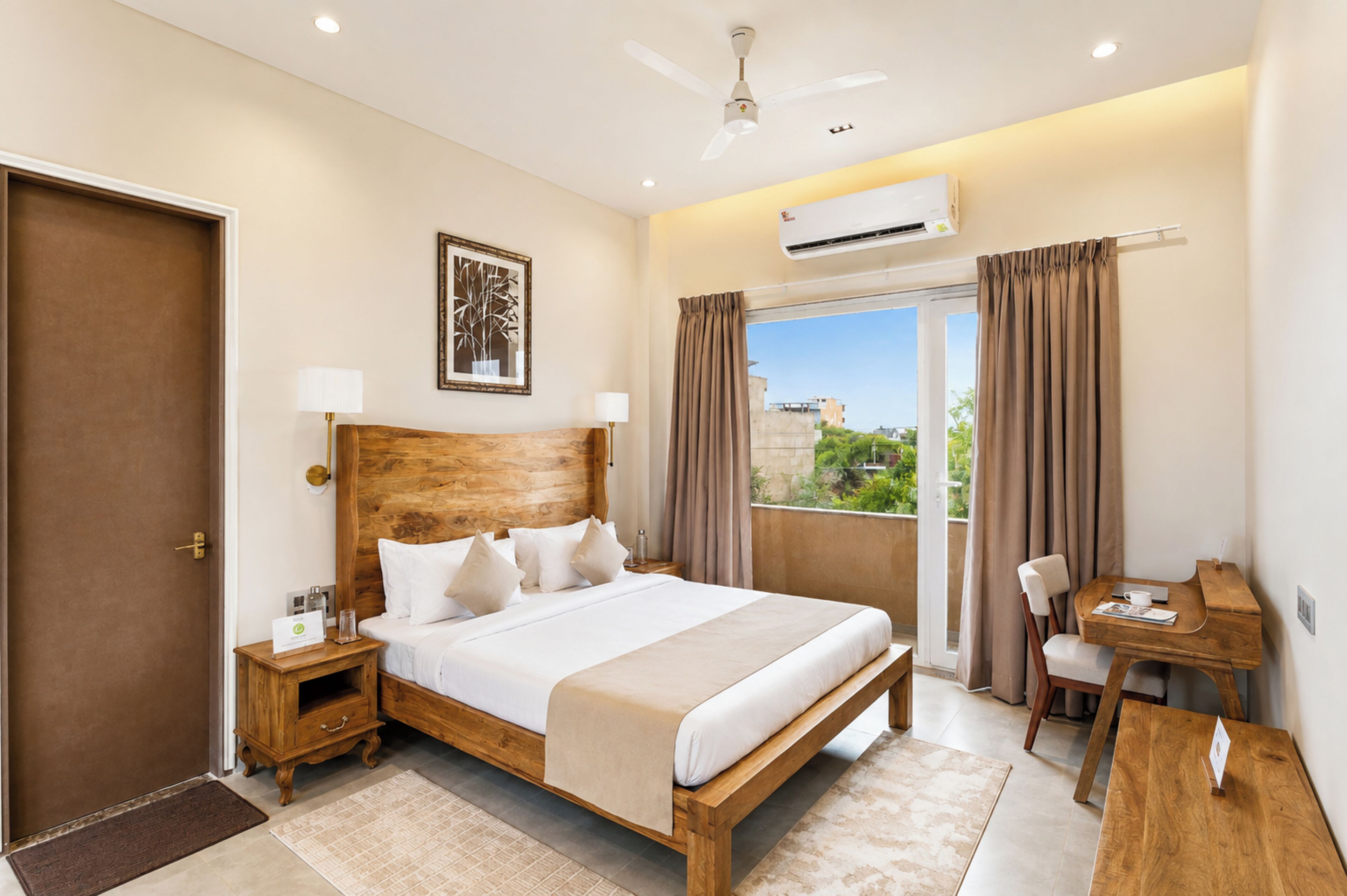 A hotel room with a wooden bed, a desk, a chair, a window with curtains, an air conditioning unit, and a door.