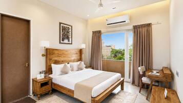 A hotel room with a wooden bed, a desk, a chair, a window with curtains, an air conditioning unit, and a door.