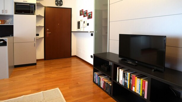 A modern living room with a flat-screen TV, a bookshelf, a microwave, and a door with a clock. - GuestHost - Good morning, Monza! Cozy Apartment (Monza)