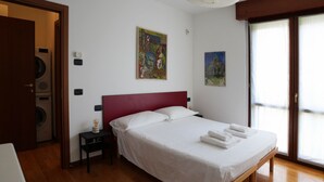 A hotel room with a bed, a nightstand, a lamp, a painting, and a window. - GuestHost - Good morning, Monza! Cozy Apartment (Monza)