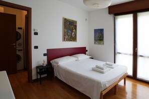 Quarto de hotel com cama, mesa de cabeceira, luminária, um quadro e uma janela.