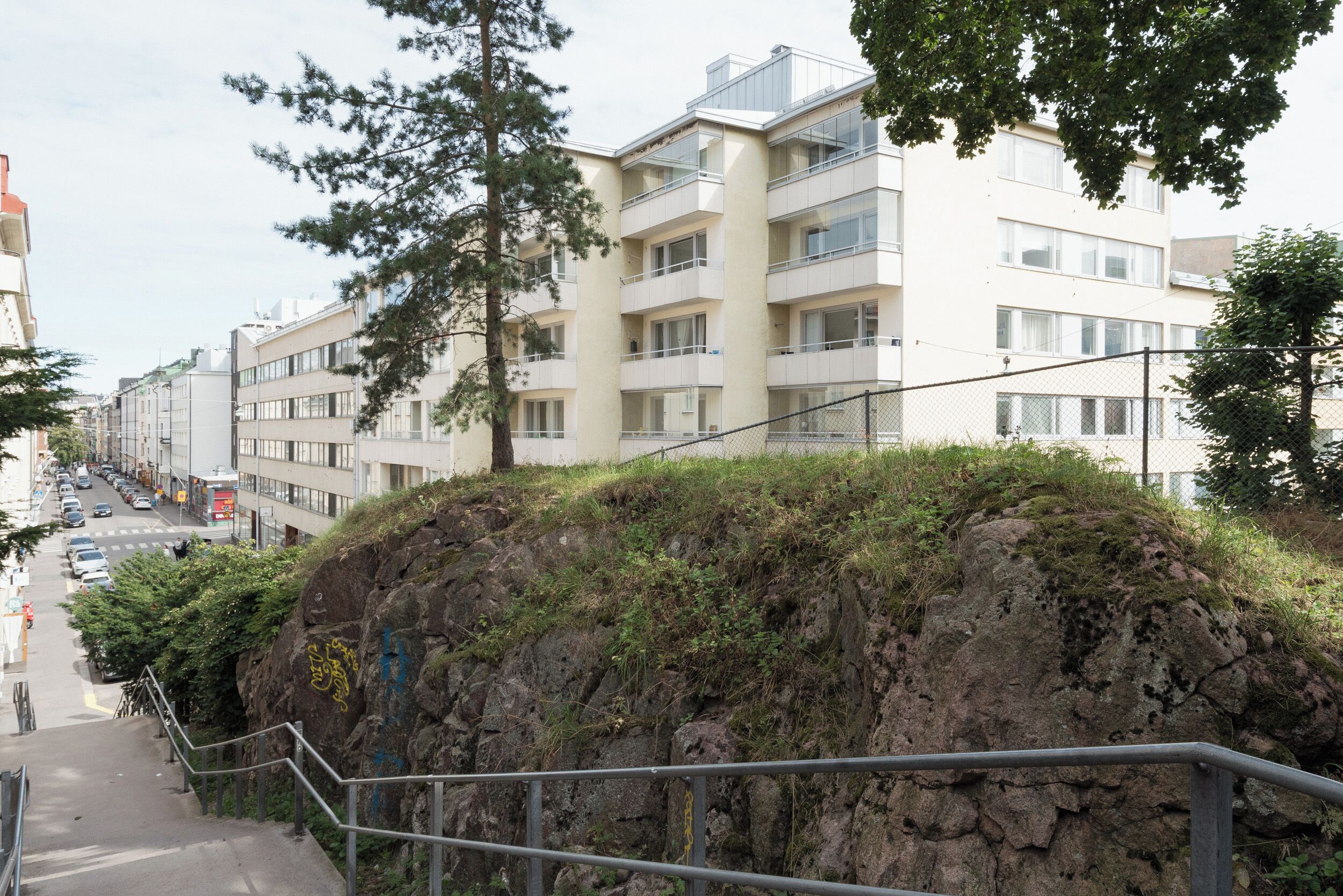 Un immeuble à plusieurs étages doté de balcons, un escalier menant à un promontoire rocheux et une rue bordée de voitures garées.