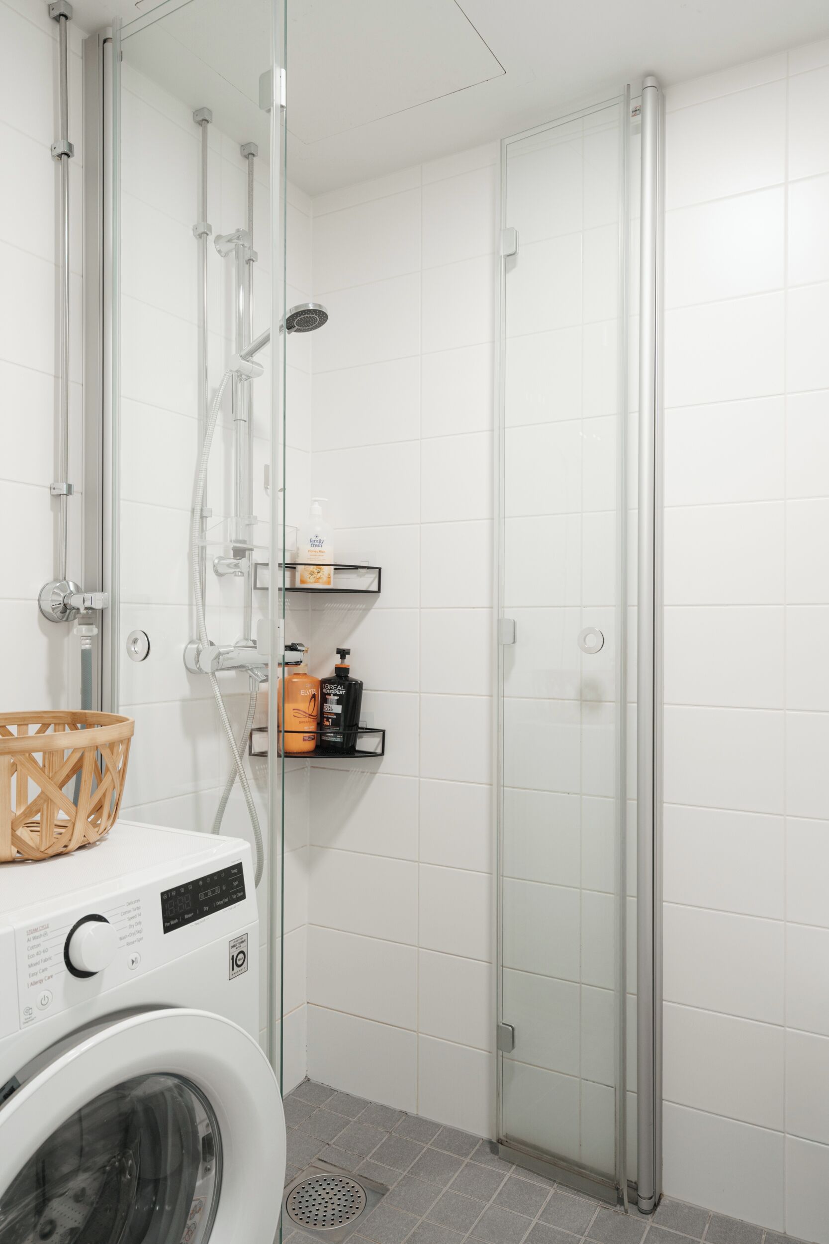 A modern bathroom with a glass shower enclosure, a washing machine, and a shelf with toiletries.
