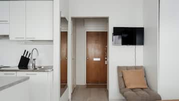 Un espace de vie compact comprenant un coin cuisine, une porte en bois, un téléviseur fixé au mur et un canapé marron avec un coussin.