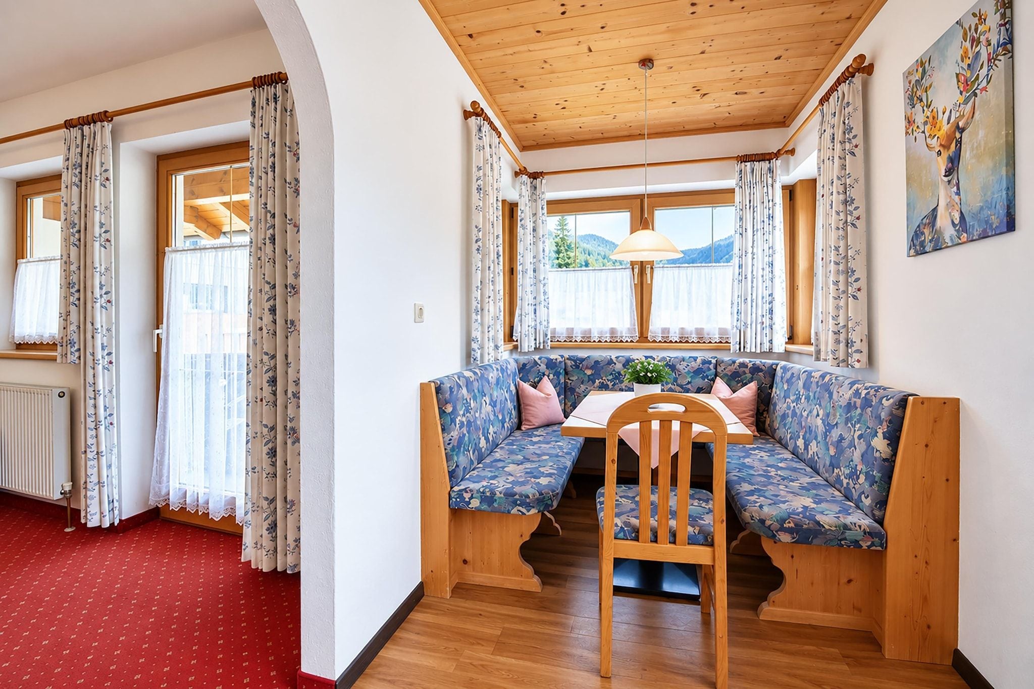 A cozy room with a wooden ceiling, a floral sofa, a wooden table, and a view of the outdoors through a window.