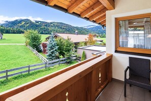 A balcony with a wooden railing, a wicker chair, and a view of a green landscape with houses and mountains. - Apartment near the lake (Achenkirch)