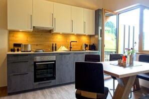 Eine moderne Küche mit eingebauten Geräten, ein Essbereich mit Holztisch und -stühlen sowie ein großes Fenster mit Blick ins Freie.