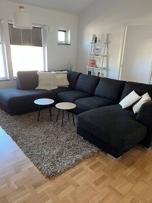 Ein modernes Wohnzimmer mit einem Ecksofa, zwei kleinen runden Tischen, einem gemusterten Teppich und einem Holzboden im Fischgrätmuster.