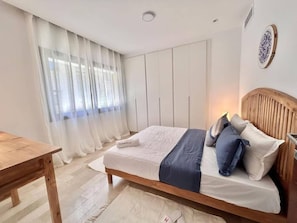 Ein Schlafzimmer mit einem hölzernen Bett, weißen Vorhängen, einem Holztisch und einer Wandleuchte.