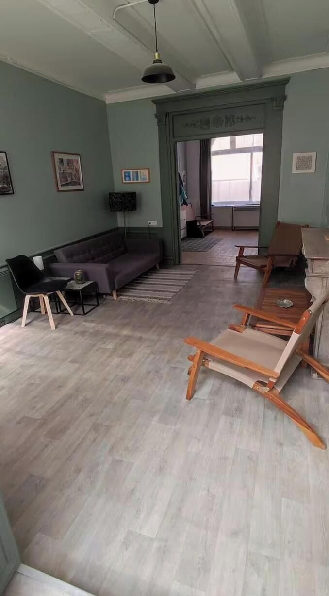 Een kamer met een licht houten vloer, een bank, een stoel en een tafel. Er is een raam met gordijnen en een plafondlamp.