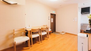 Ein Zimmer mit einem Holztisch, vier Stühlen, einer kleinen Mülltonne, einem weißen Gerät und einem Fernseher an der Wand.