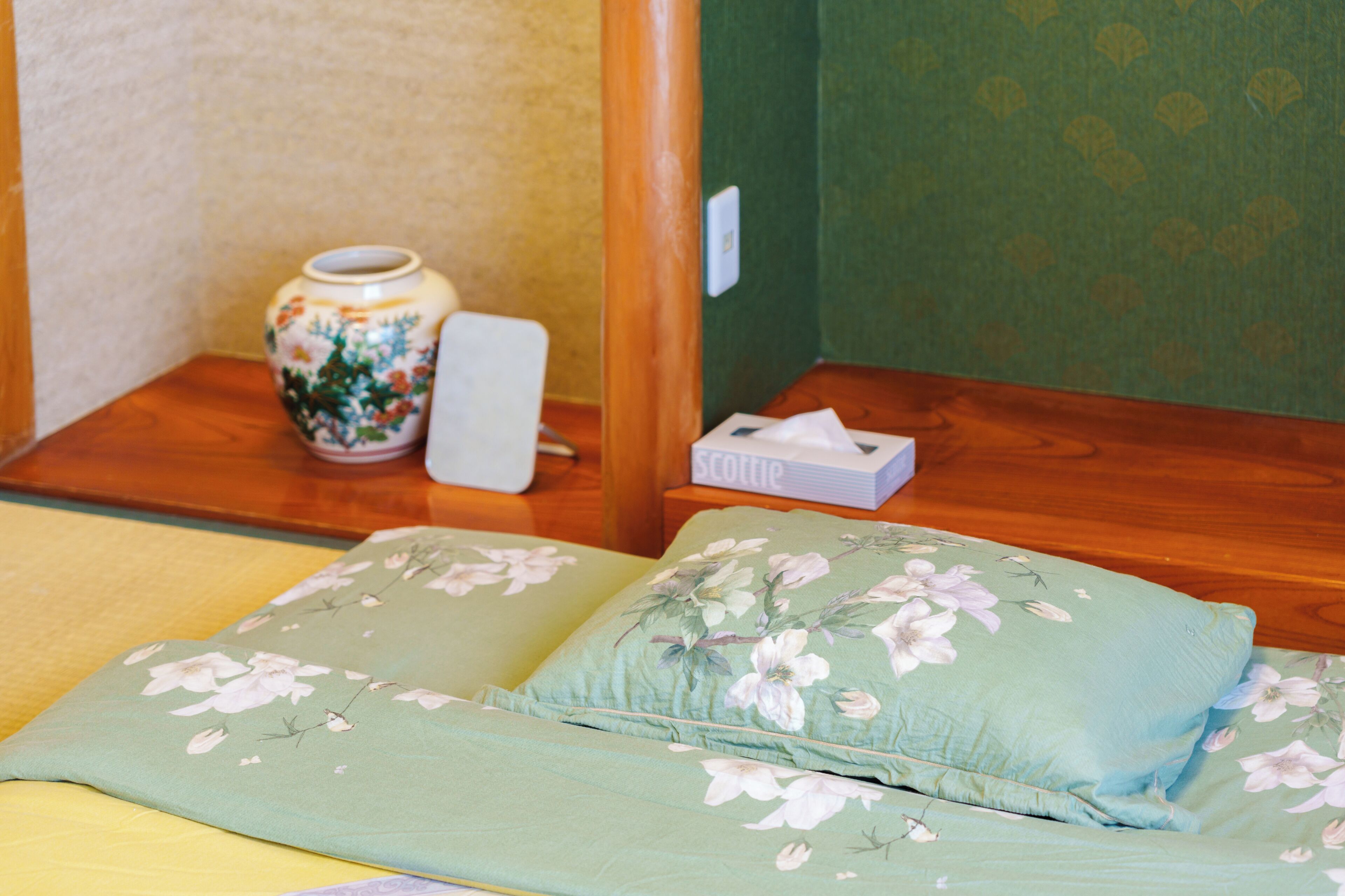 A room with a wooden floor, a green patterned wall, a floral pillow, a white vase, and a tissue box.