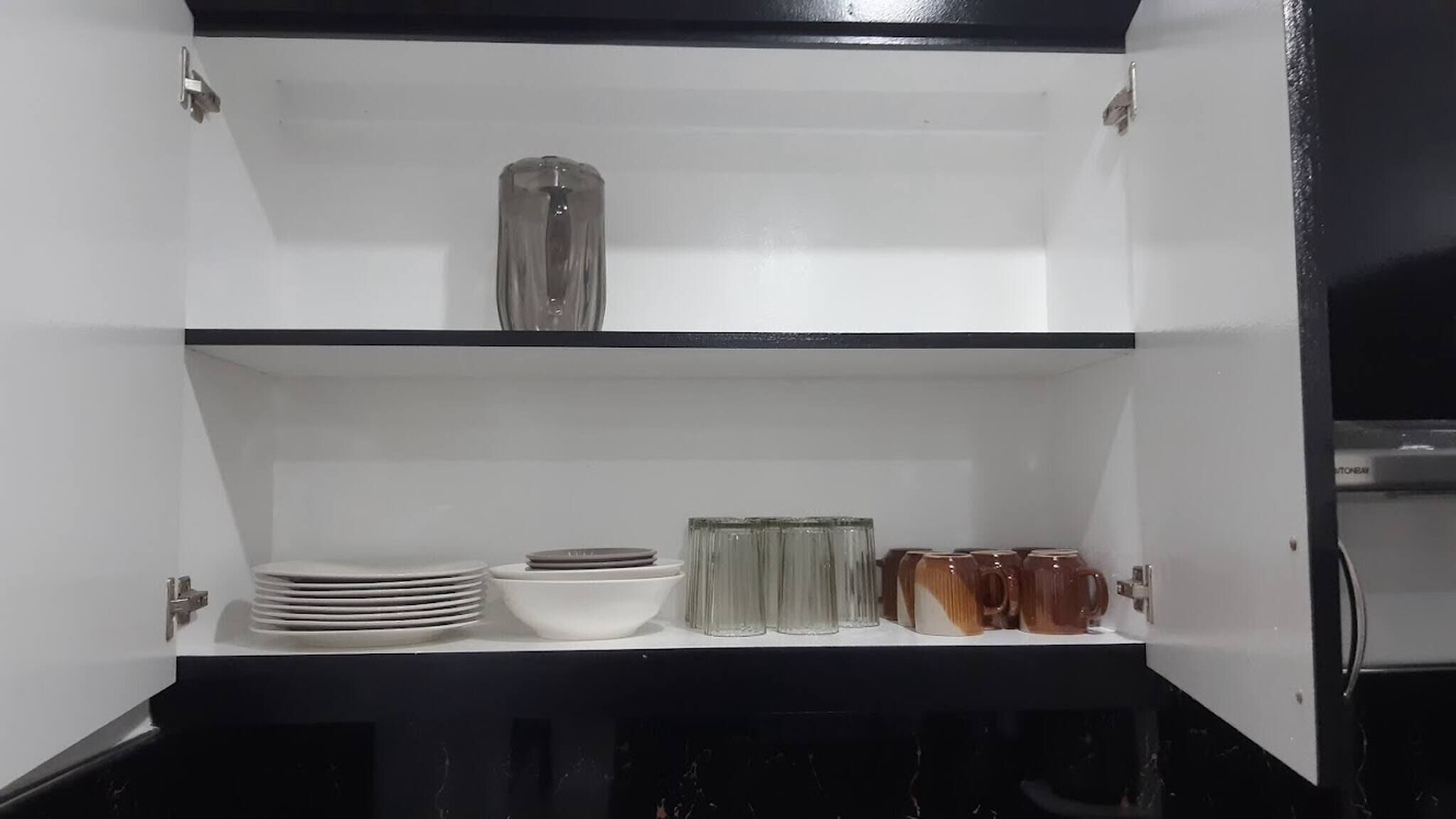 Un armario blanco con estantes que contienen platos, tazones, vasos y una jarra.