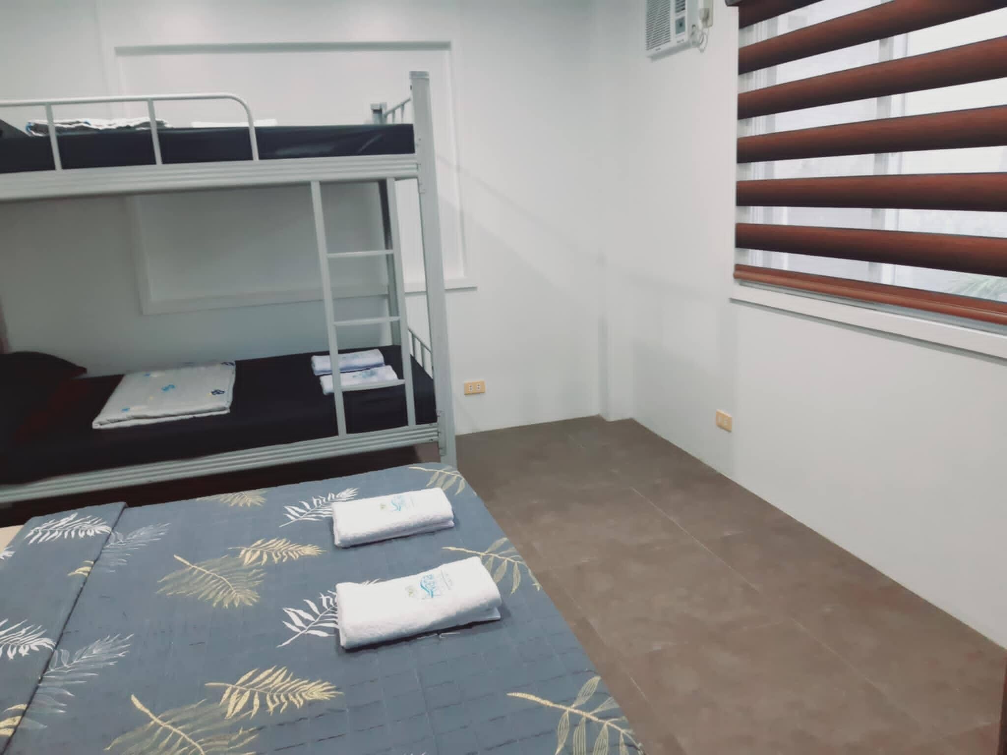 A dormitory room with bunk beds, a window with blinds, and a bed with a leaf-patterned bedspread.