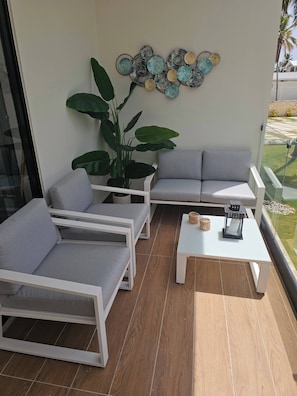 A balcony with a grey sofa, white armchairs, a potted plant, and a decorative wall piece. - Eden Tropical new 1BR Condo, Walk to Beach ,Pool (Punta Cana)