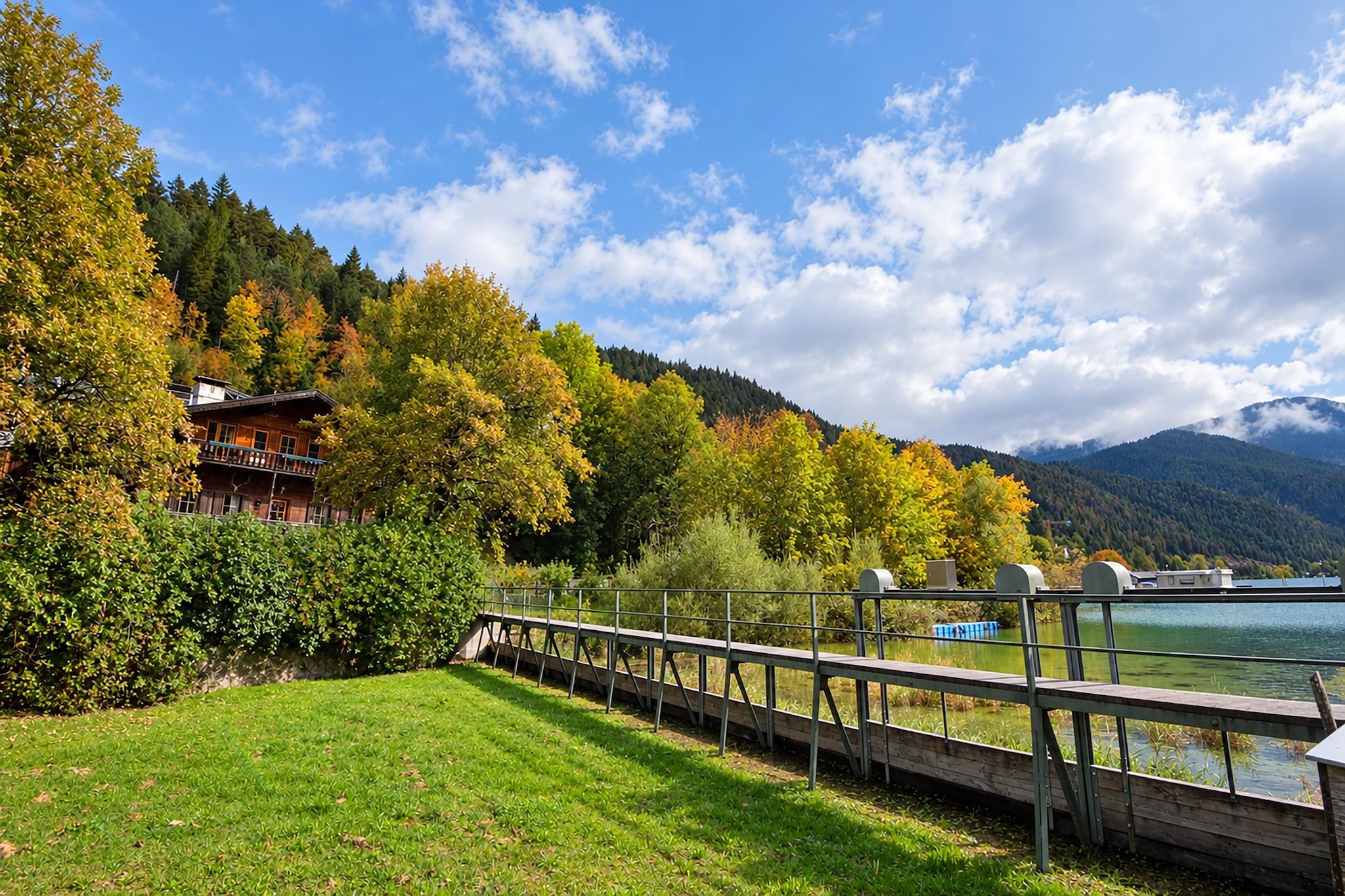 A lakeside property with a wooden deck, a building with a balcony, and a mountain backdrop.