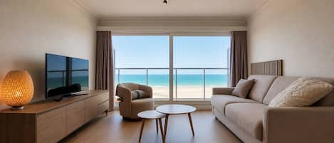 A modern hotel room with a large window offering a beach view, a sofa, a coffee table, and a TV on a wooden console.