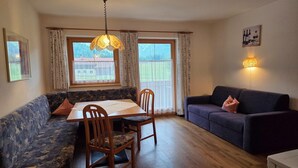 Una sala acogedora con una mesa de comedor, sillas de madera, un sofá y un sillón. Hay dos ventanas con cortinas, una lámpara colgante y un cuadro en la pared.